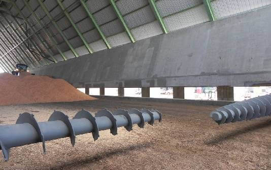 Interior of an industrial facility featuring large augers on the ground, with piles of material and a high, sloped ceiling.