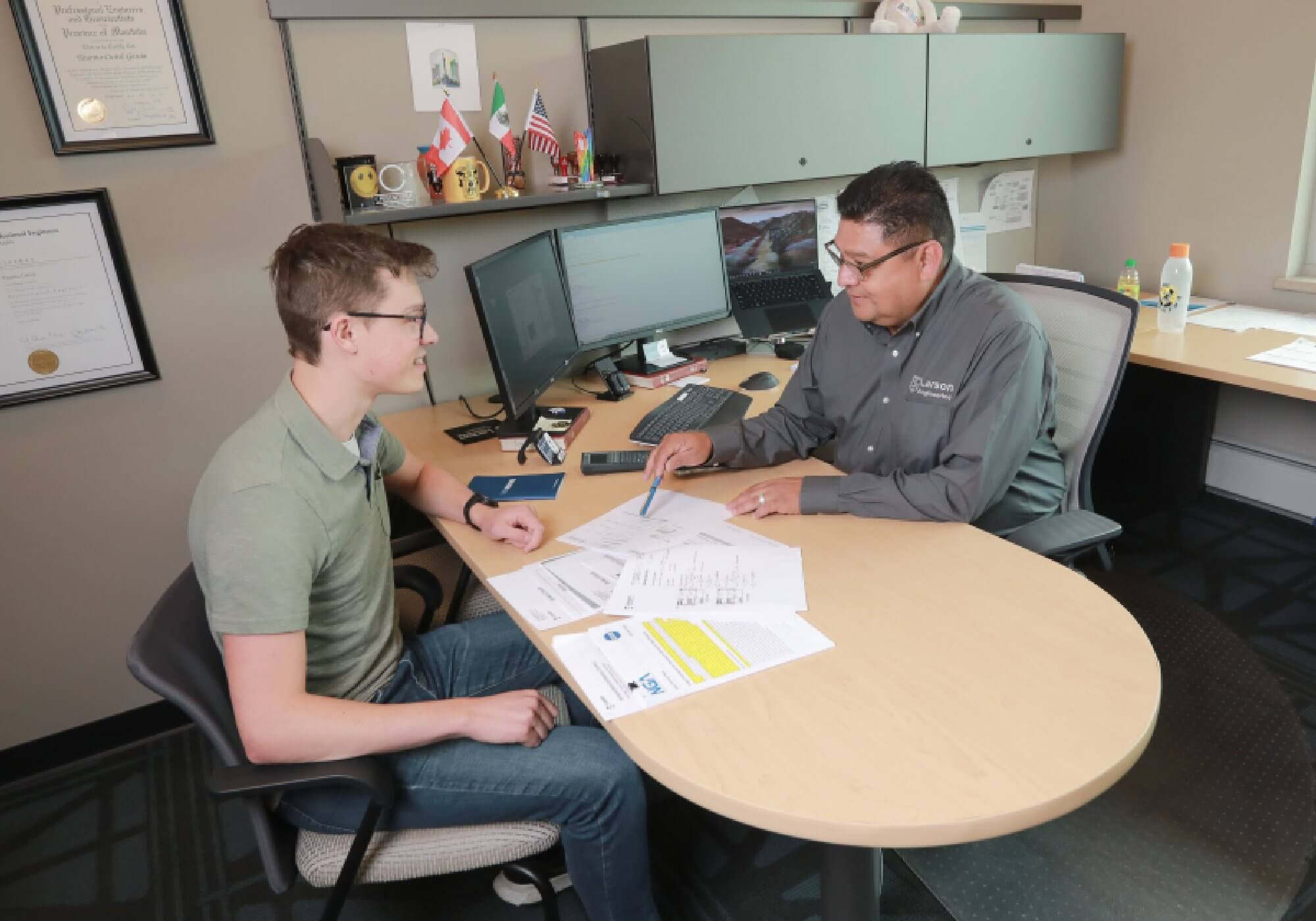 Engineer discussing project plans with a client in an office, with documents and computer screens visible on the desk.