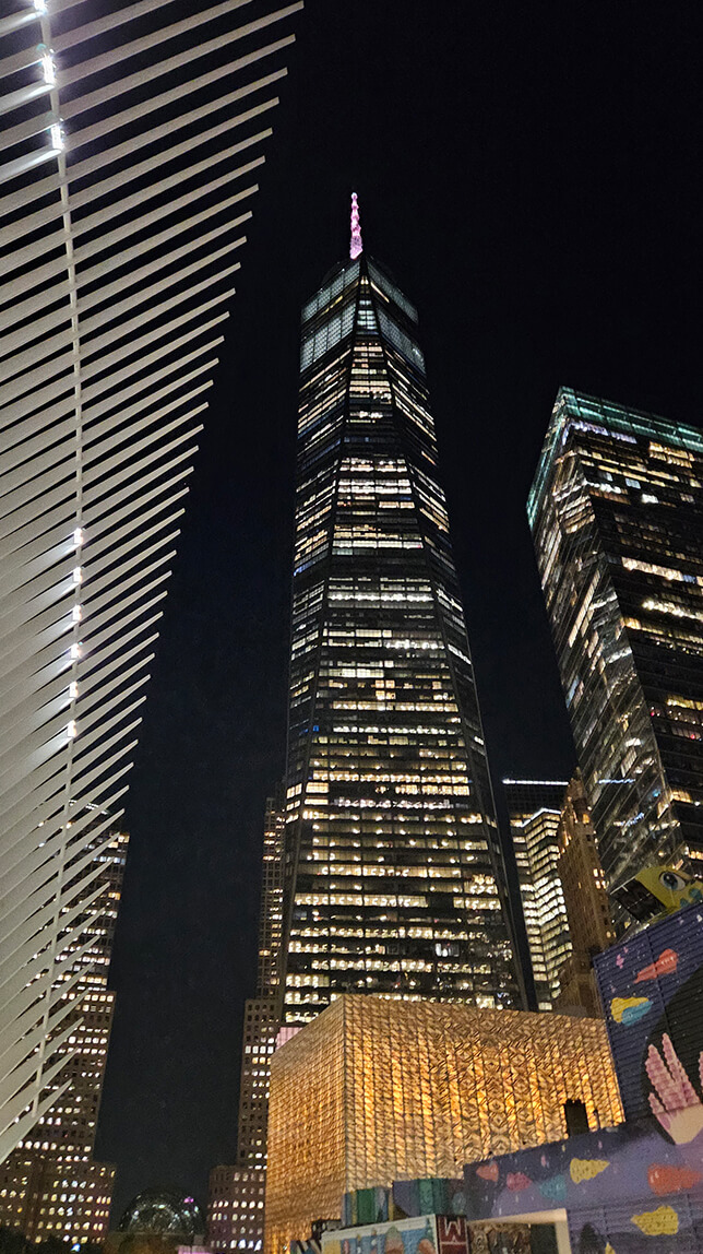 Tall skyscraper illuminated at night, surrounded by other buildings, with a modern architectural structure in the foreground.