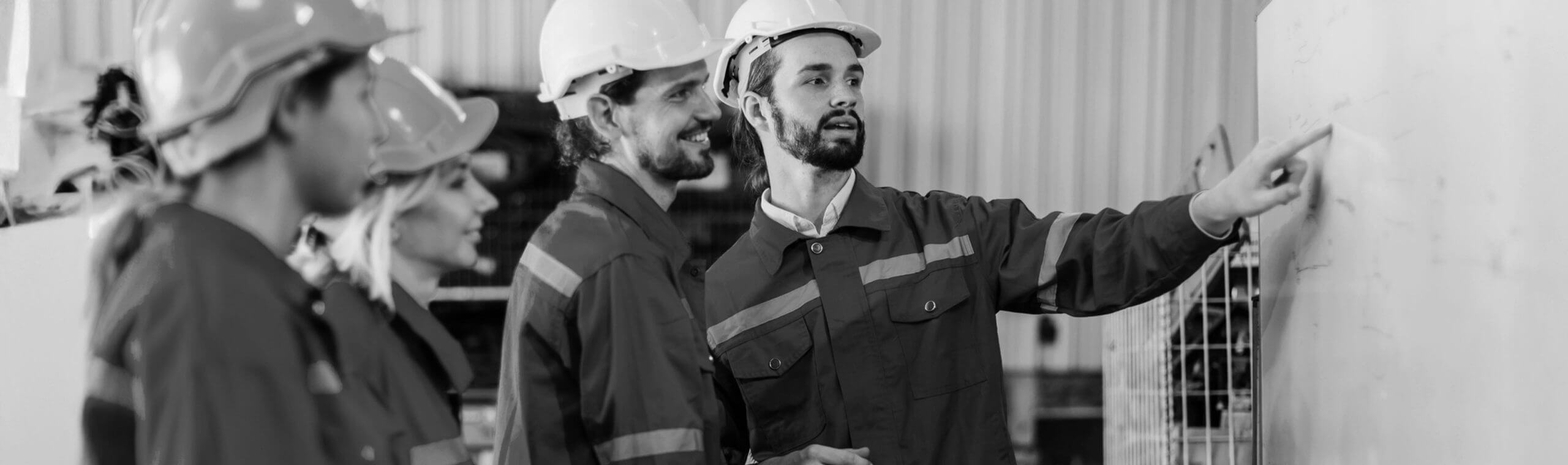 Engineers wearing hard hats and uniforms discuss a project while one points at a large structure, illustrating collaboration in an industrial setting.