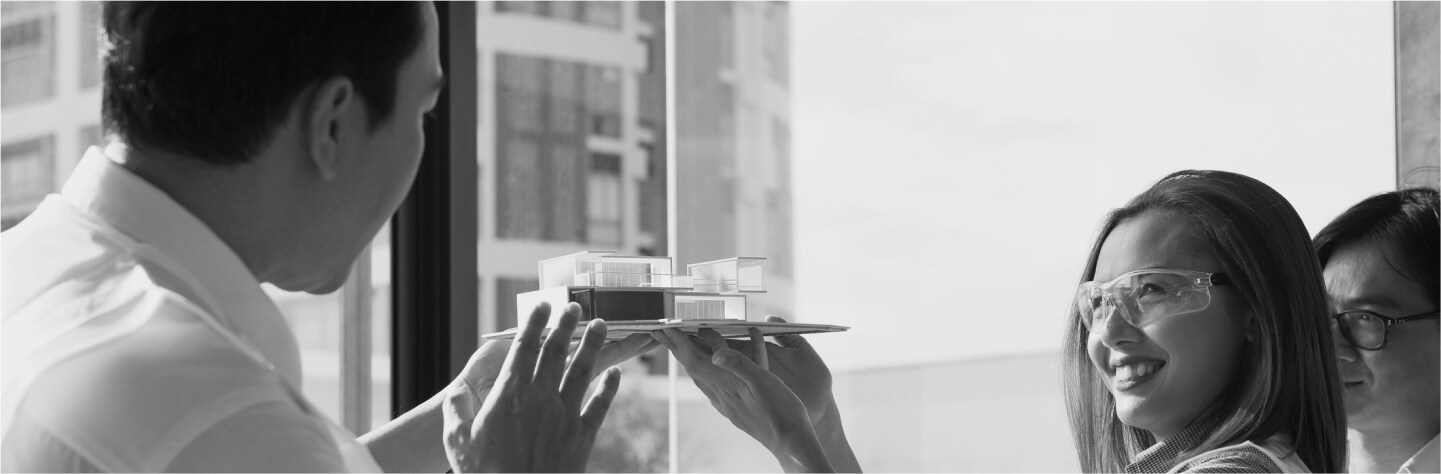 Architects discussing a building model in an office setting, with one person holding the model and another smiling while wearing safety glasses.