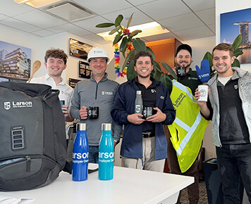 Larson Engineering employees smiling and holding branded mugs and bottles in an office setting, with a backpack and safety vest visible on a table.