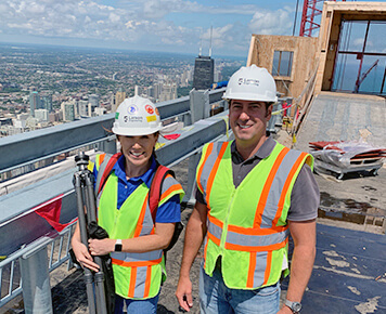 Two construction professionals wearing safety vests and helmets stand on a high-rise building under construction, overlooking a cityscape.
