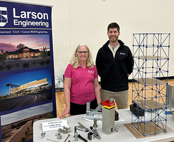 Larson Engineering representatives at a career fair booth displaying engineering models and promotional materials, with a banner featuring the company name and images of their projects.