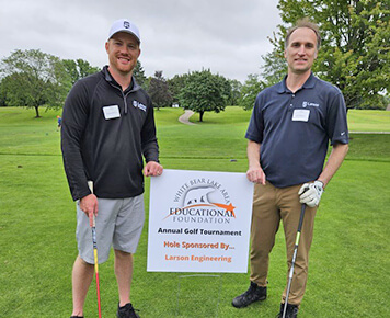 Two people standing on a golf course holding clubs, smiling next to a sign that reads, "White Bear Lake Area Educational Foundation Annual Golf Tournament. Hole Sponsored By... Larson Engineering."