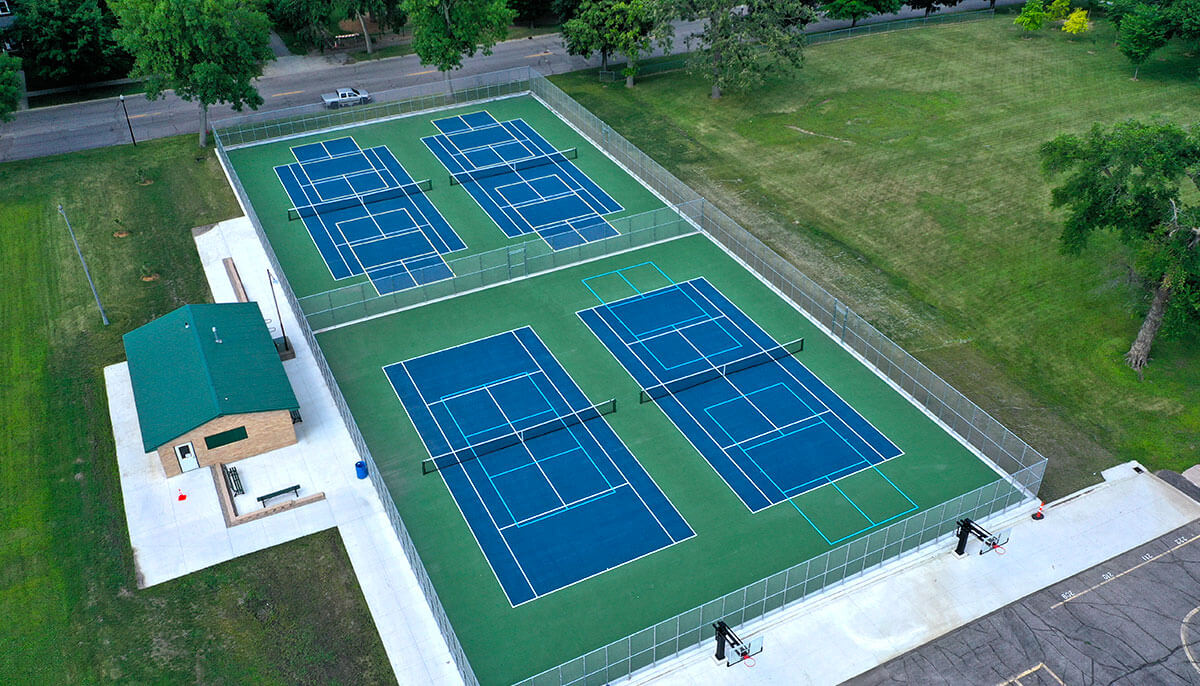 Aerial view of a tennis court complex with six blue courts surrounded by fencing, adjacent to a small green-roofed building, set in a grassy area with trees nearby.