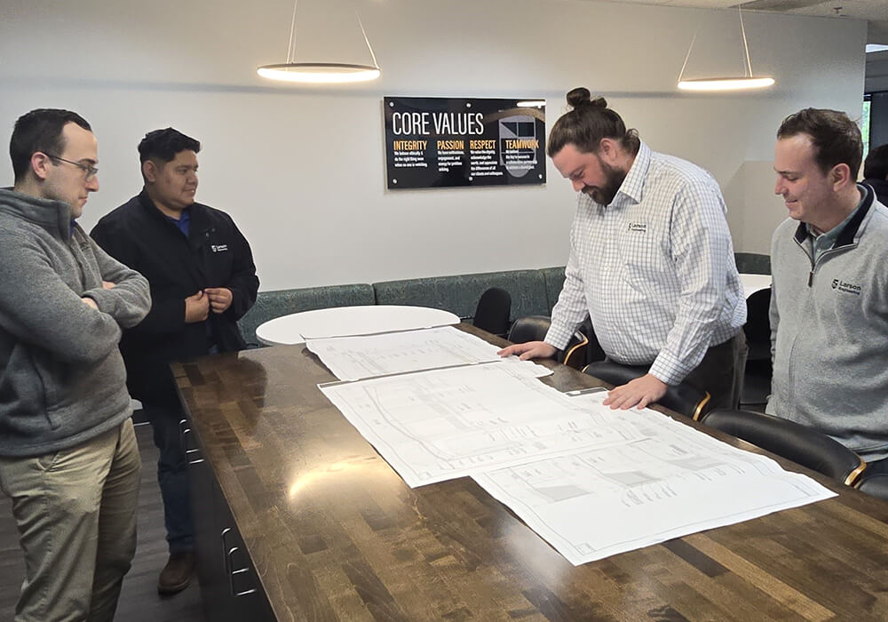 Engineers reviewing large architectural blueprints on a conference table, with a "Core Values" sign displaying "Integrity, Passion, Respect, Teamwork" on the wall behind them.