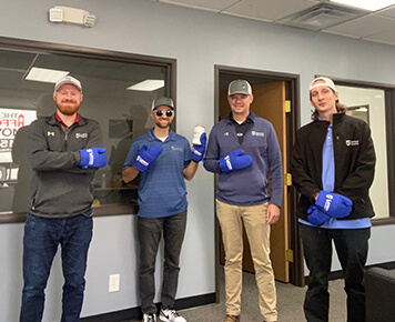 Four Larson Engineering employees stand indoors, smiling and wearing branded hats and blue mittens, reflecting a casual and friendly workplace culture.