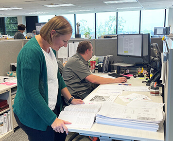 Employee reviewing documents at a desk in an open office environment, with a colleague working on a computer in the background.