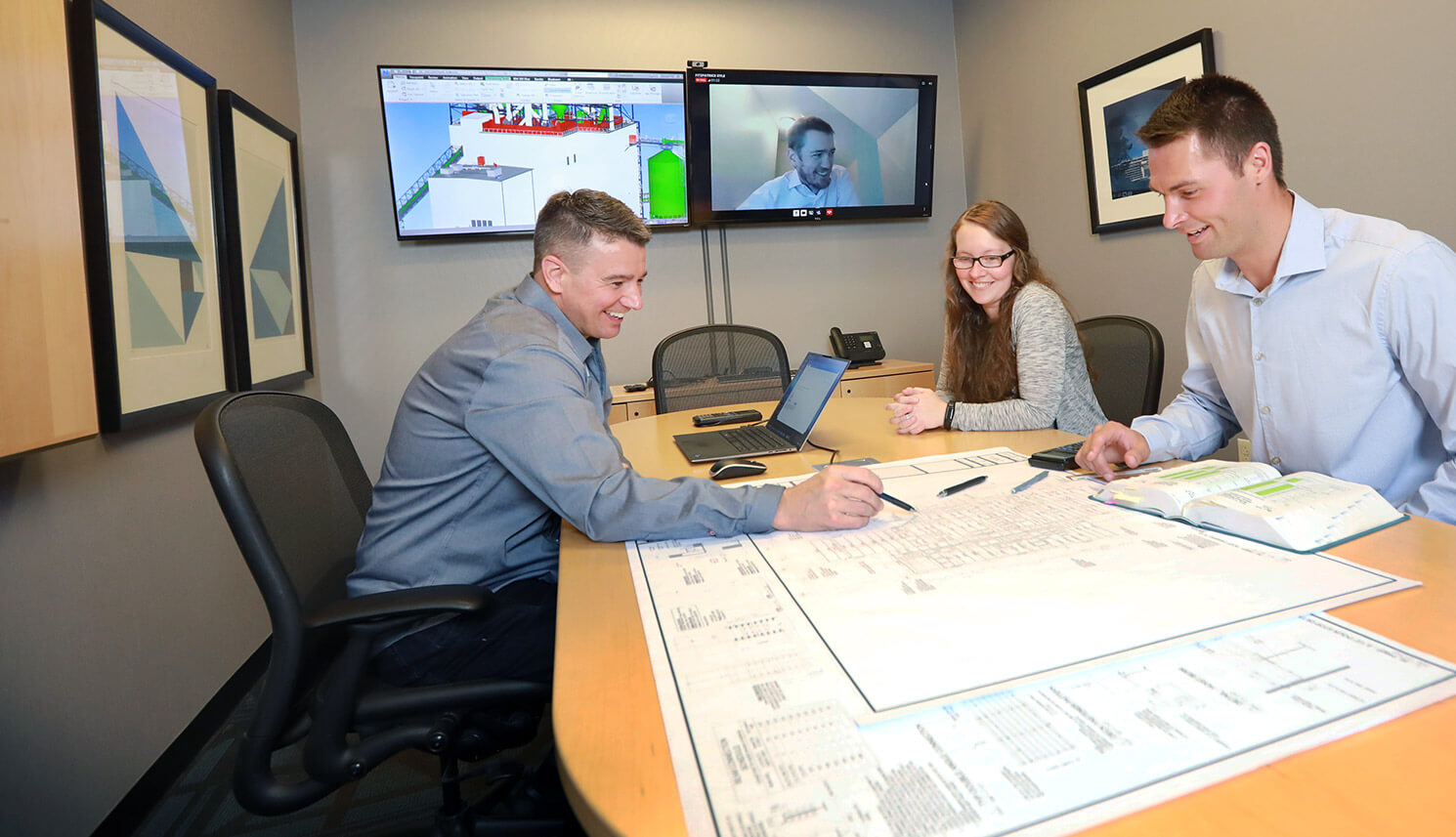Engineers collaborating around a conference table with architectural plans, two screens displaying design software, and a video call in progress.
