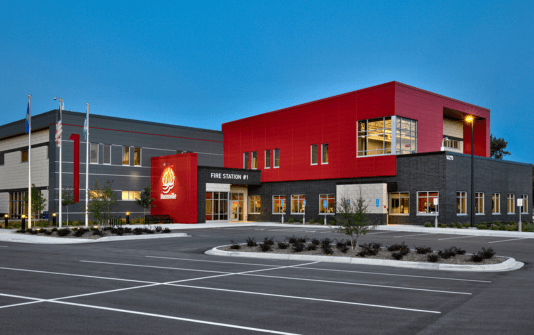 Modern fire station building with a red and gray exterior, labeled "FIRE STATION #1" and "Blaine" on the facade, surrounded by a parking lot and landscaping.