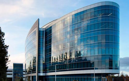 Modern glass façade of a curved office building reflecting the sky and surrounding trees, illustrating architectural design expertise.