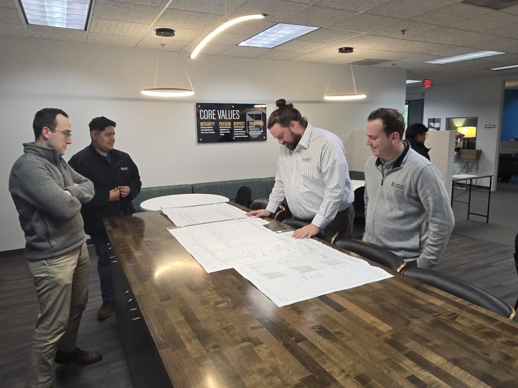 Engineers reviewing architectural blueprints on a conference table in an office, with a "Core Values" sign on the wall.