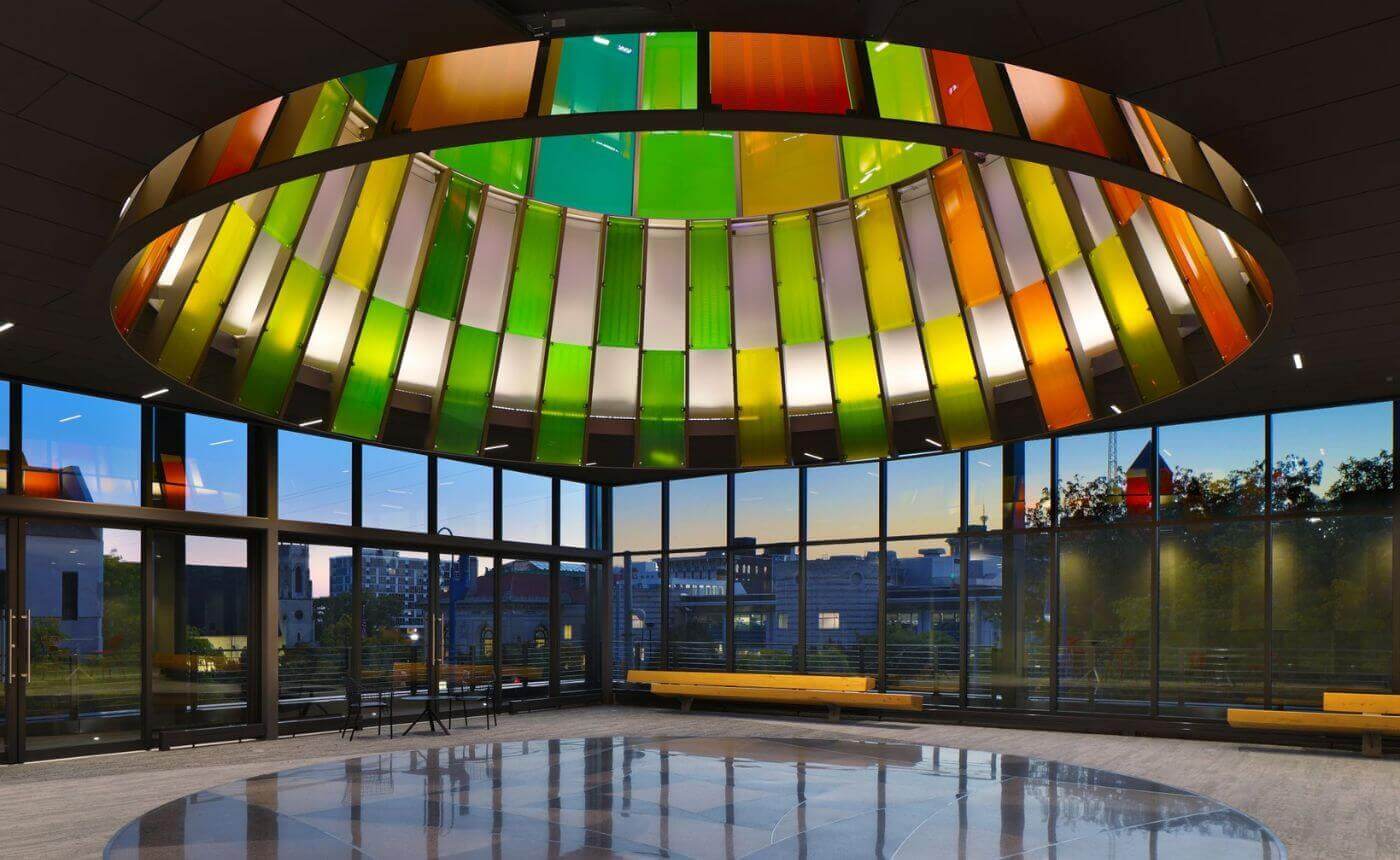 Colorful skylight with green, yellow, and orange panels illuminating a modern, circular lobby with large windows and a reflective floor at dusk.