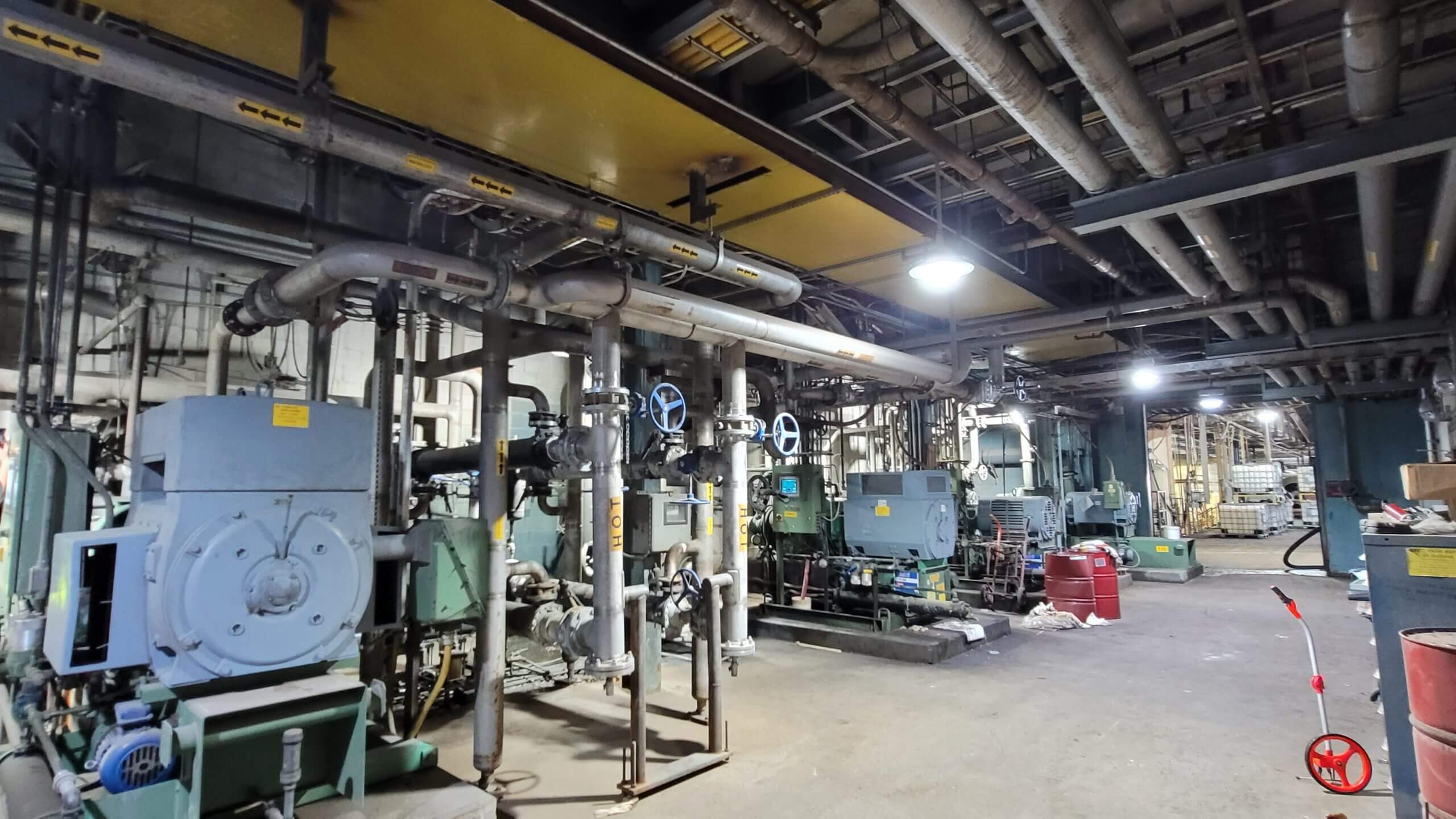 Industrial mechanical engineering facility with complex piping, machinery, and equipment under a network of overhead pipes and ducts.