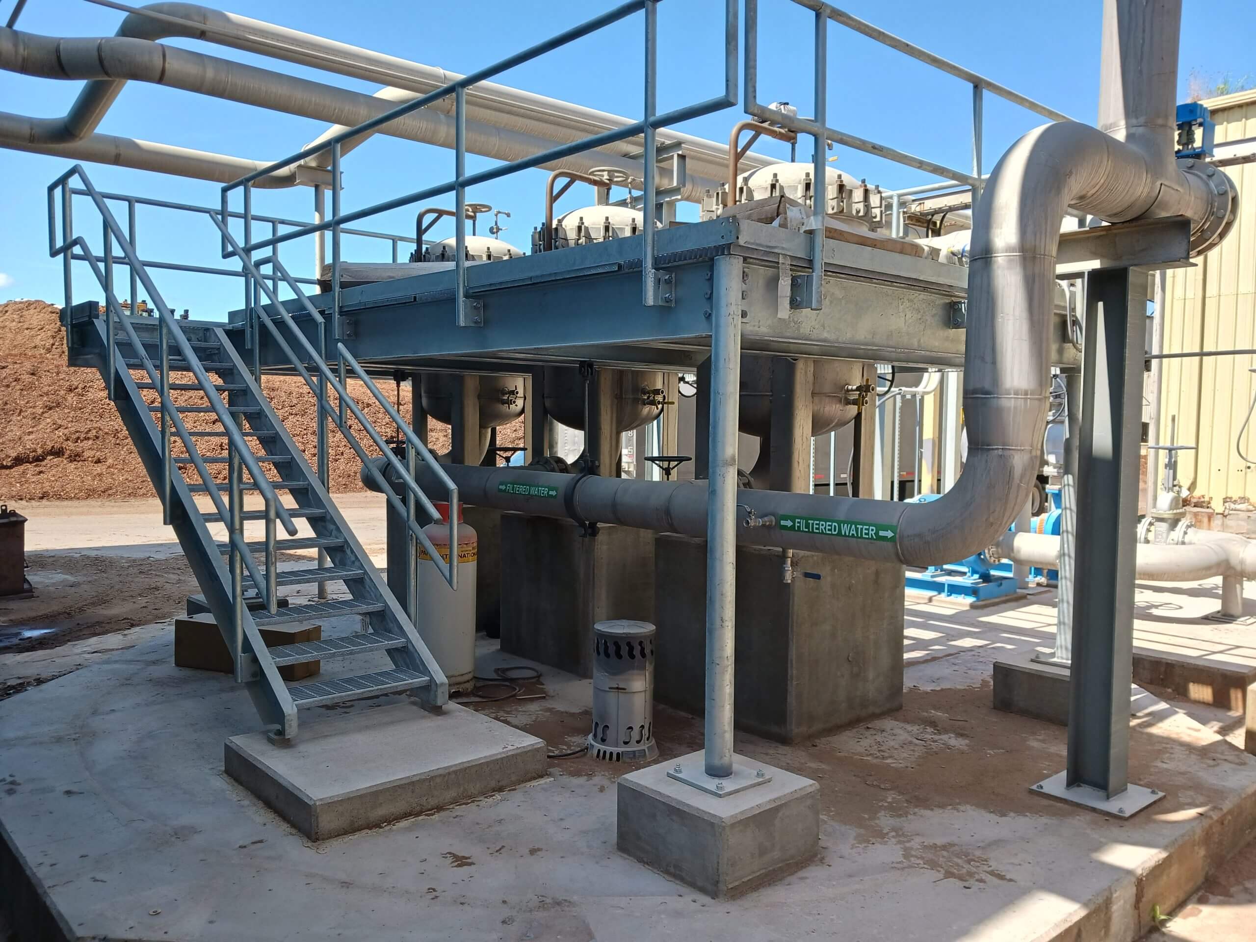 Industrial piping system with elevated metal platform, stairs, and labeled pipes, including one marked "FILTERED WATER," situated outdoors under a clear sky.