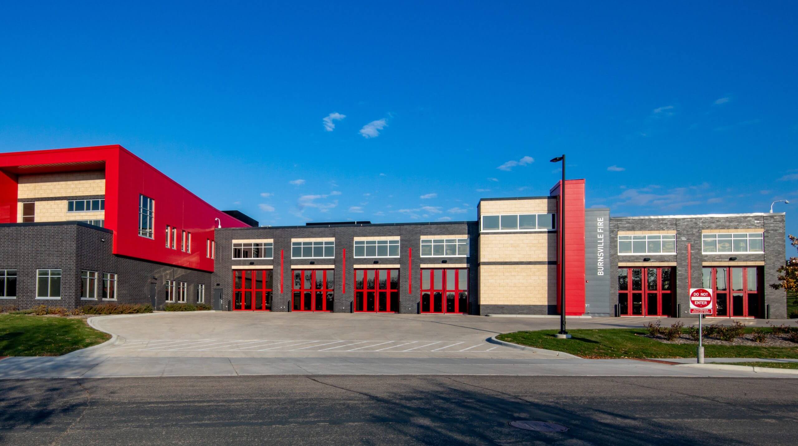 Modern building with a mix of red, beige, and gray brick, featuring large windows and red garage doors, set against a clear blue sky.