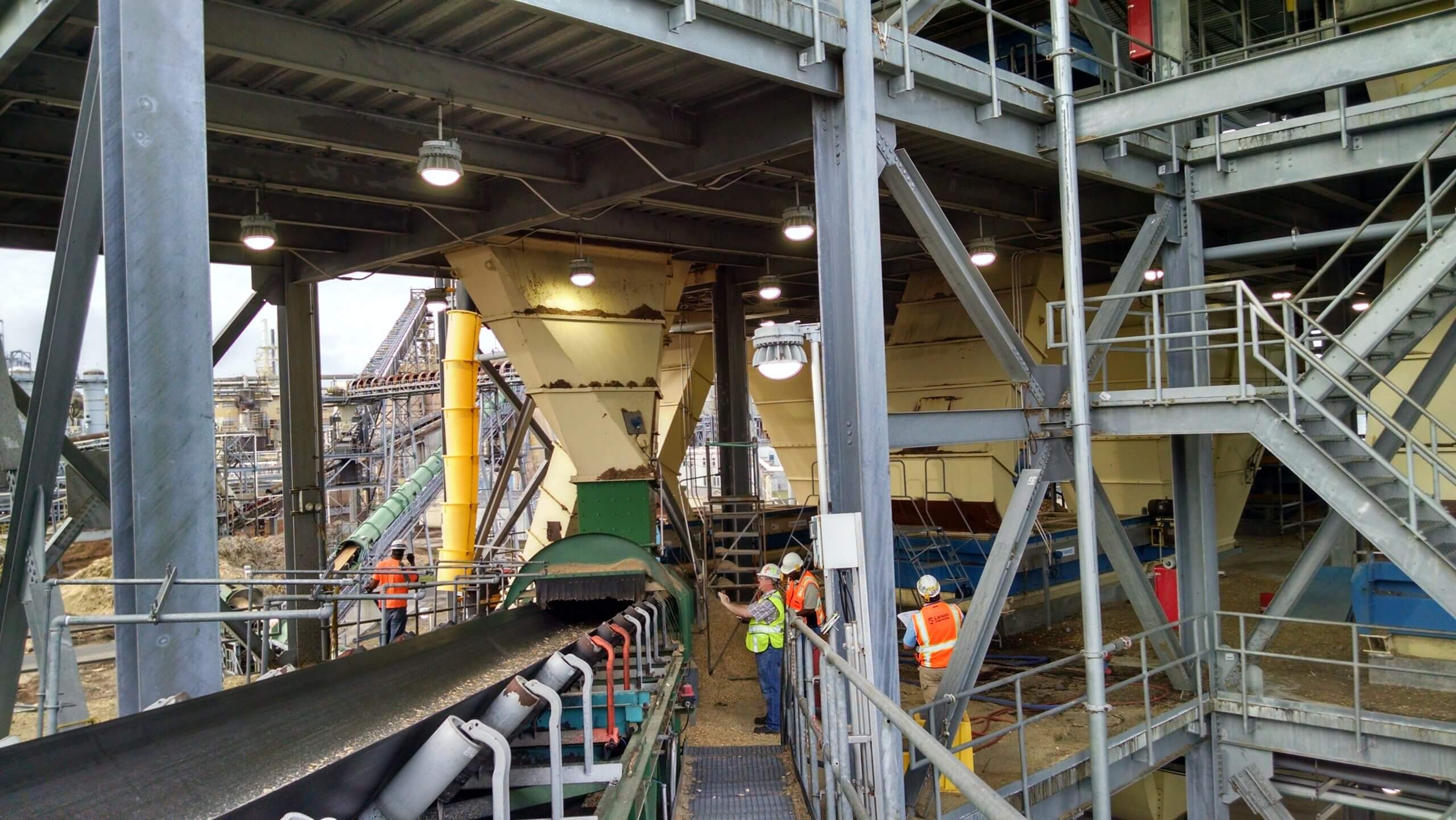 Industrial facility with engineers in safety vests and helmets inspecting machinery and conveyor systems under a large metal structure.