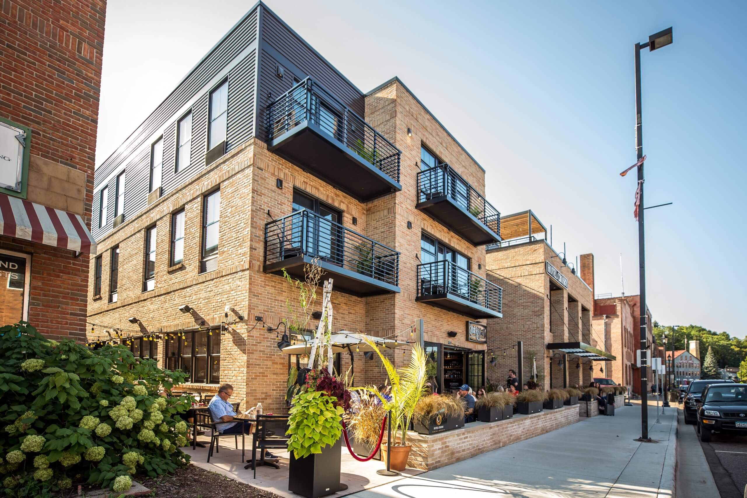Modern brick building with balconies and outdoor seating, located on a city street with pedestrians and plants nearby.