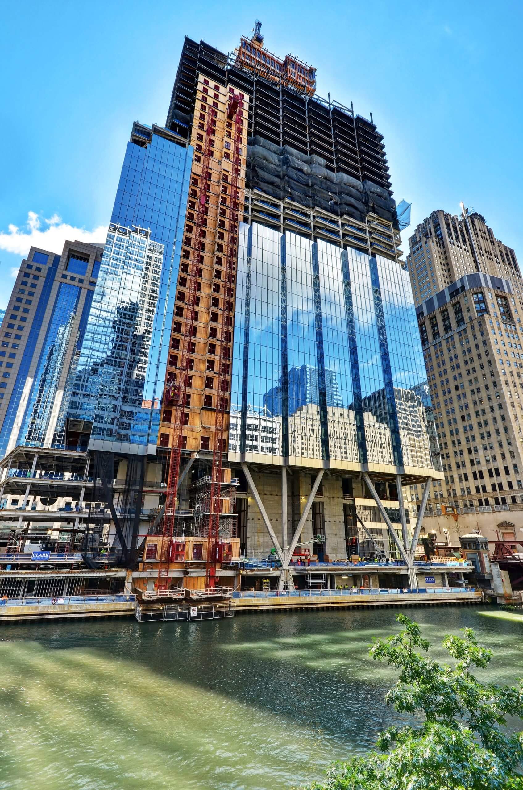 St. Regis Chicago under construction, featuring a prominent construction hoist on the building's side, with reflective glass windows and a view of the river in the foreground.