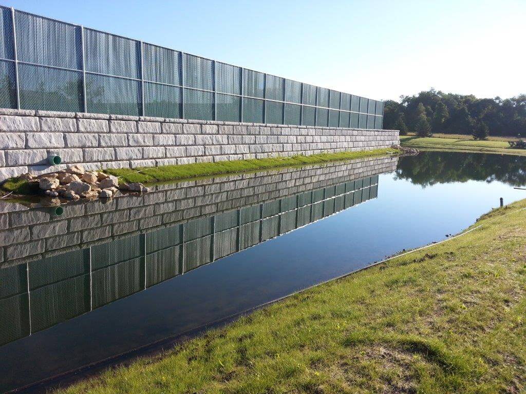 Stormwater retention pond with a stone retaining wall and metal fence, reflecting the clear sky and surrounding greenery.