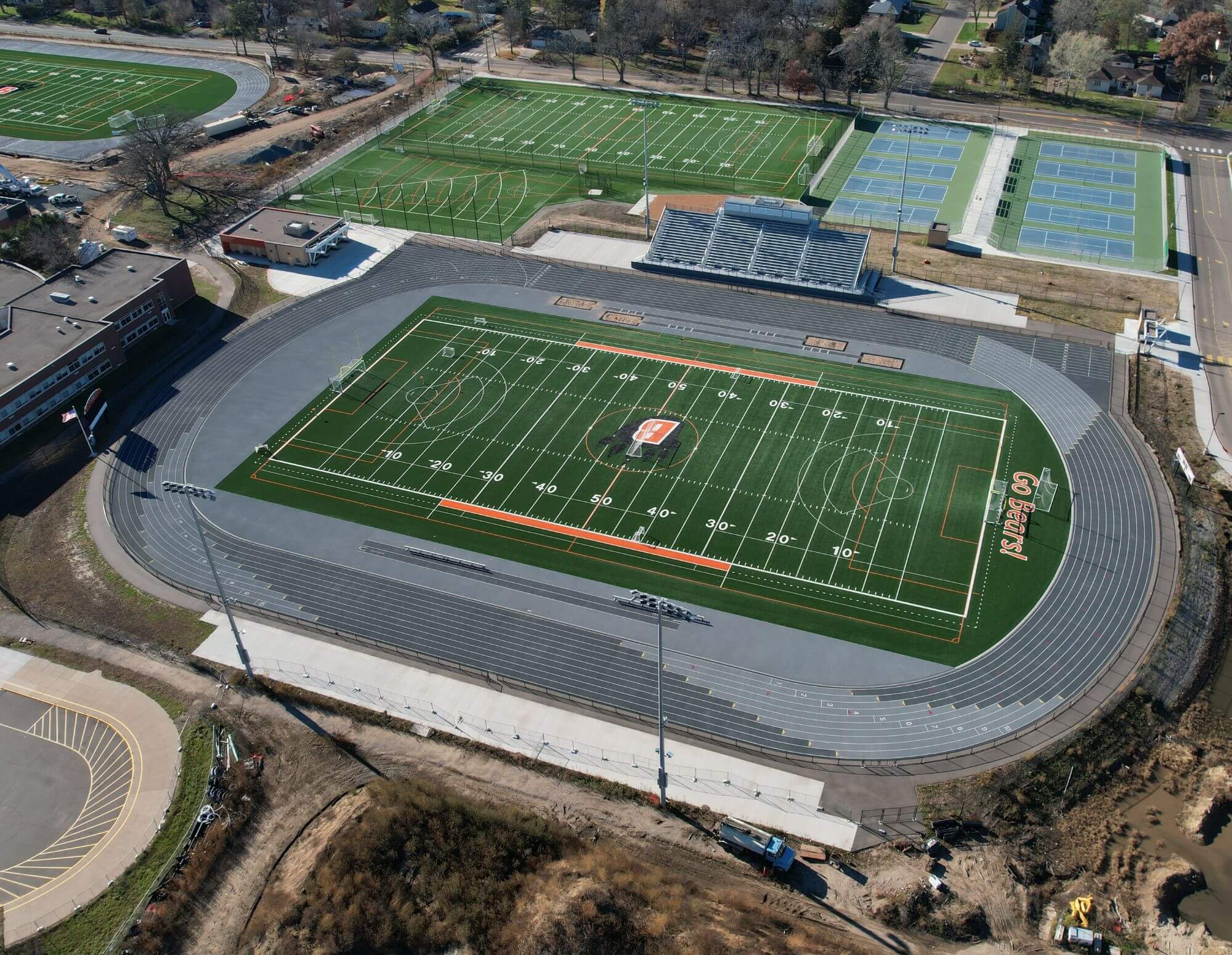 Aerial view of athletic facilities featuring a large football field with a surrounding track, multiple tennis courts, and additional sports fields in the background.