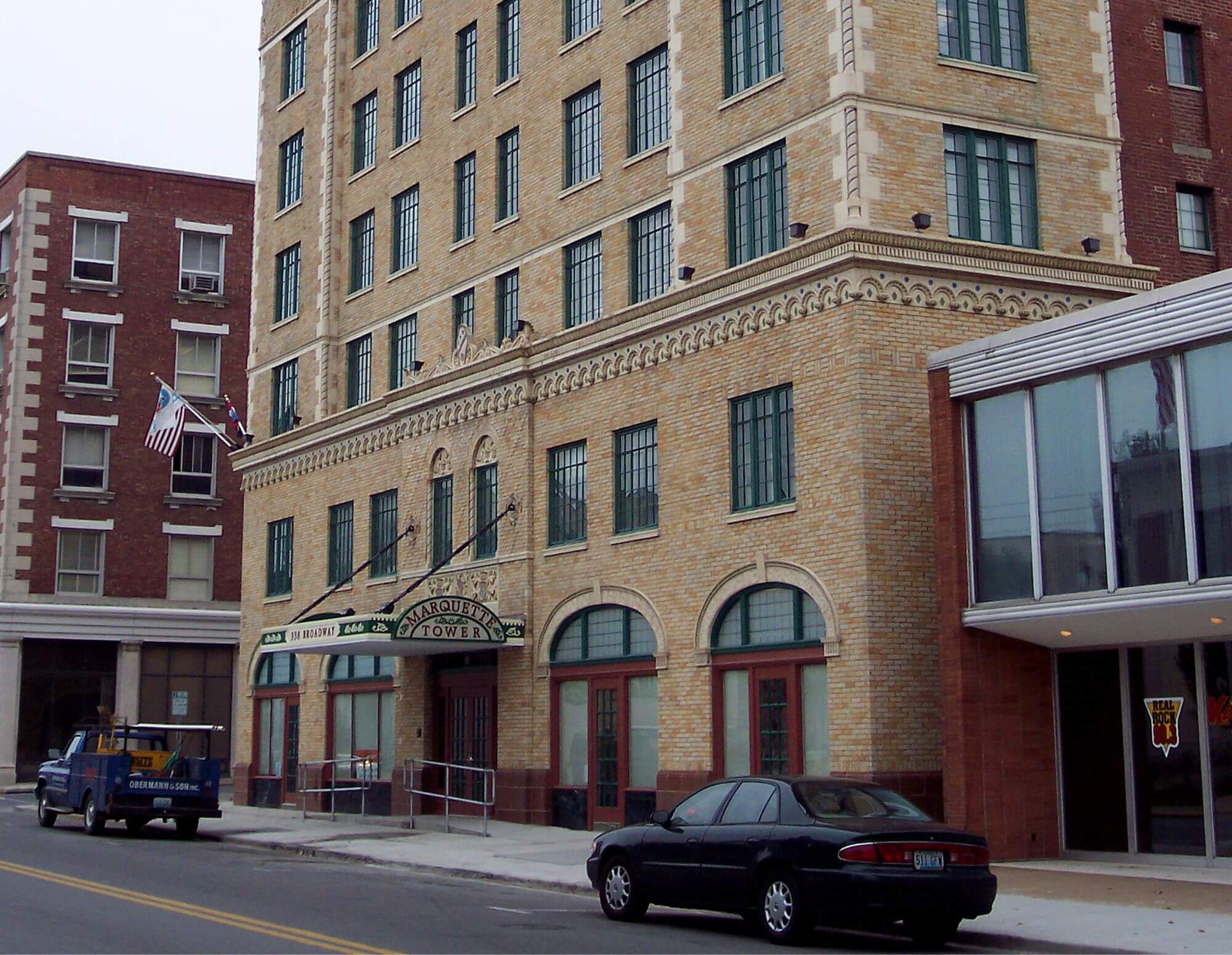 Historic multi-story brick building with arched windows and a sign reading "Larson's Tower" above the entrance, flanked by parked cars and an American flag.