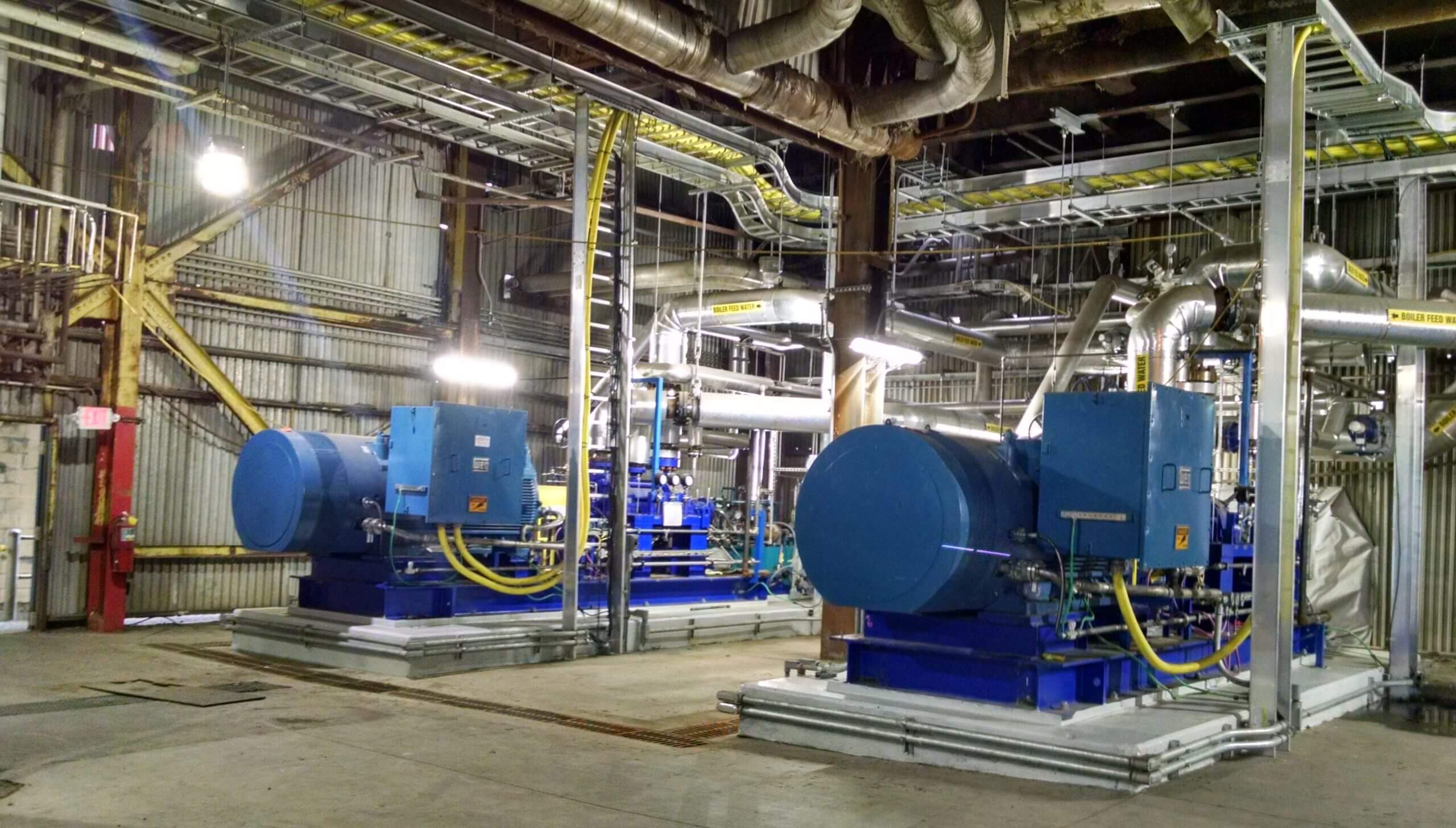 Industrial facility interior featuring two large blue boiler feed pumps connected by yellow hoses, surrounded by metal pipes and overhead ductwork.
