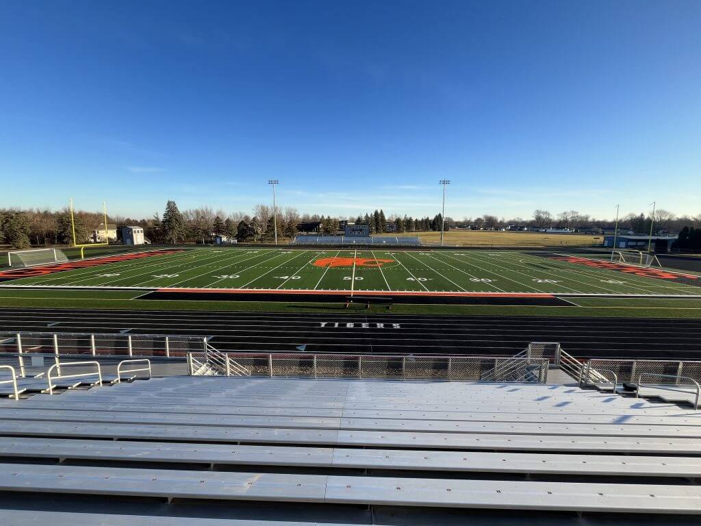 Football field with a large orange "G" at the center, surrounded by bleachers and goalposts, under a clear blue sky.