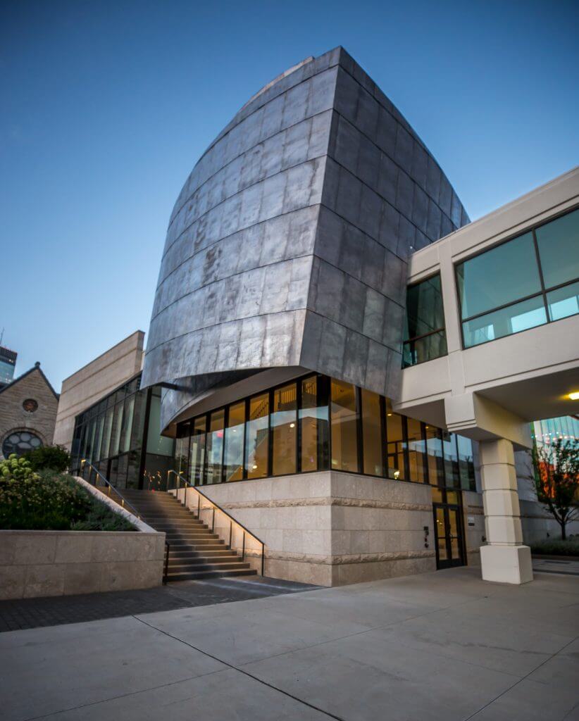 Modern building with a unique curved metal facade and large glass windows, featuring an exterior staircase leading to the entrance.