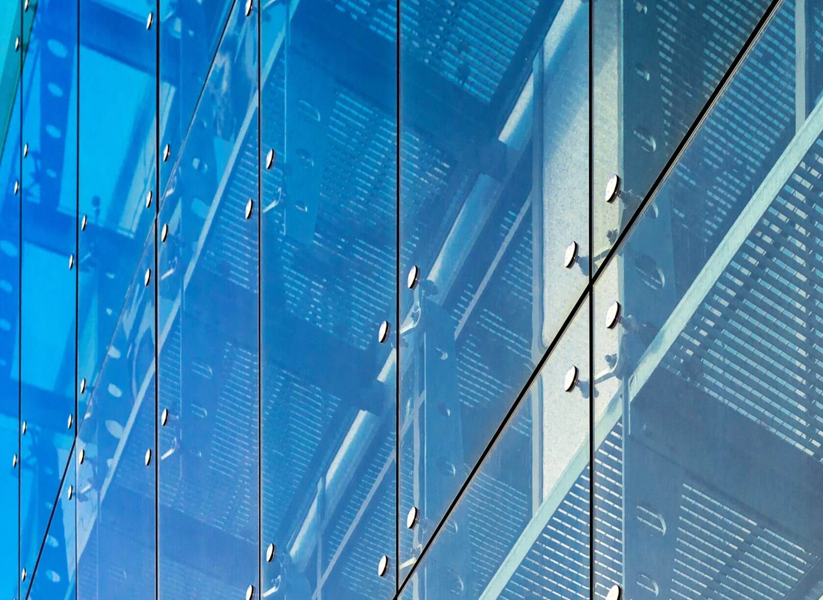 Glass facade of a modern building reflecting structural elements and a blue sky, showcasing architectural precision and design.