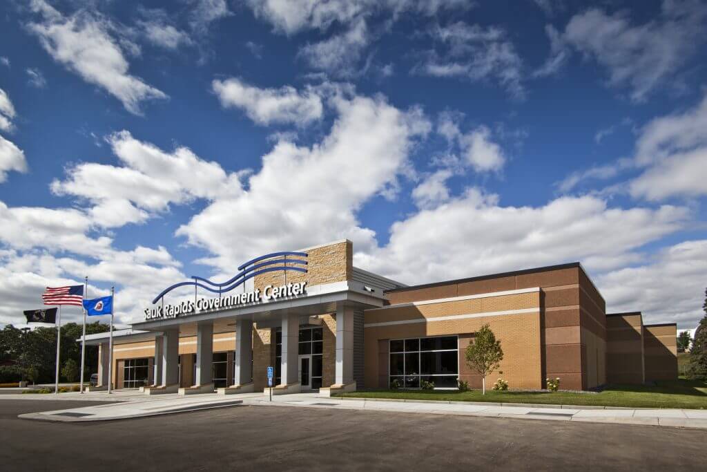 Sauk Rapids Government Center building under a partly cloudy sky, featuring a modern brick facade and the text "Sauk Rapids Government Center" prominently displayed above the entrance.