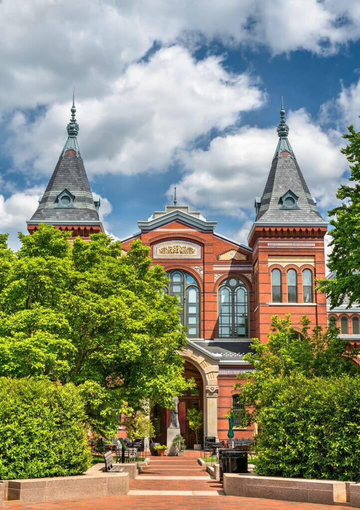 Arts and Industries Building of the Smithsonian museums in Washington, D.C., featuring red brick architecture, two spired towers, and lush greenery in the foreground.