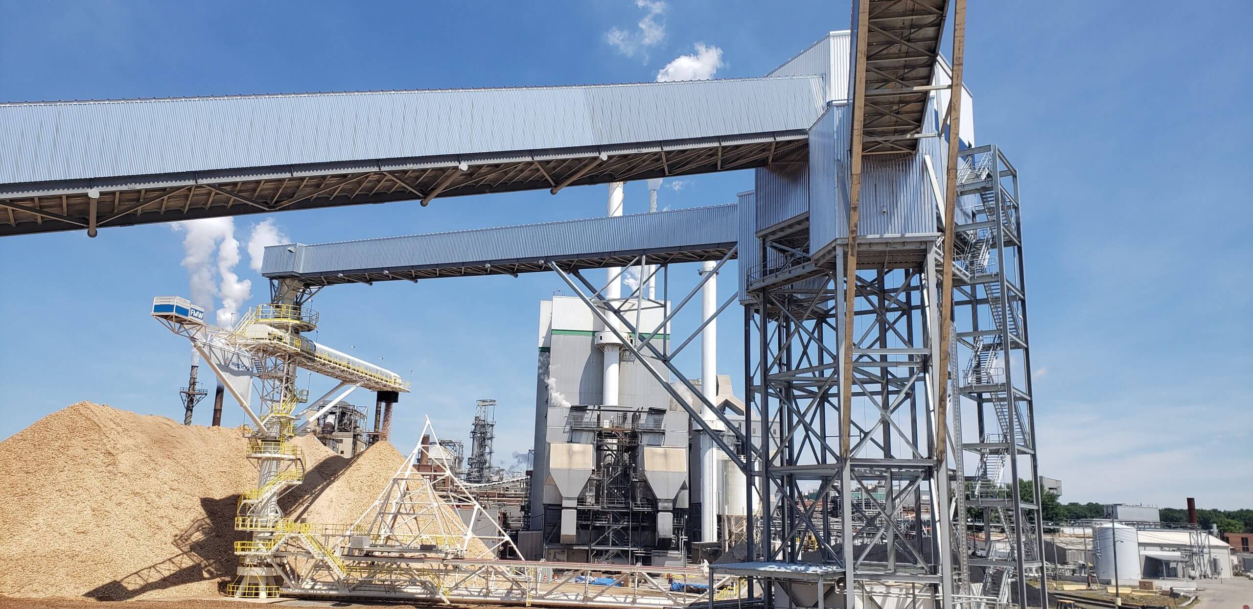 Industrial facility with large metal structures and conveyors, piles of wood chips, and a clear blue sky.