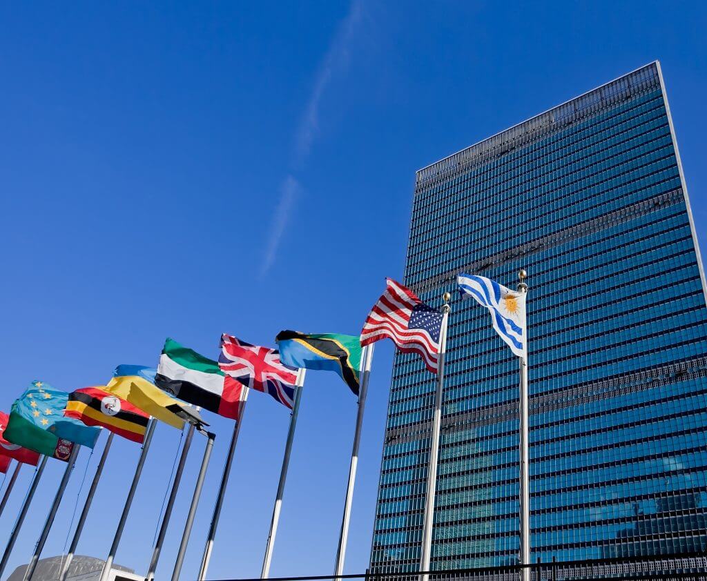 Tall modern glass building with multiple international flags, including the United States and United Kingdom, against a clear blue sky.