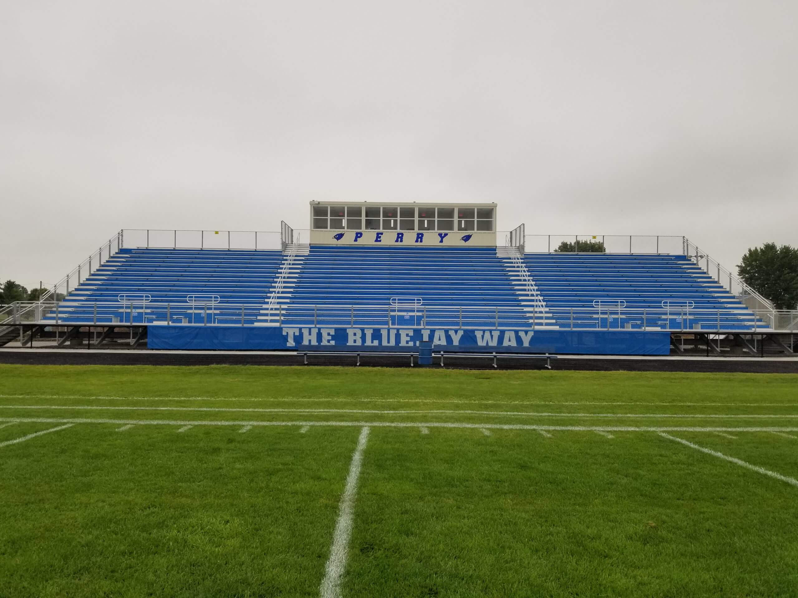 Stadium bleachers with blue seats and a press box above, featuring the text "PERRY" and "THE BLUEJAY WAY" at Perry Community School District athletic facility.