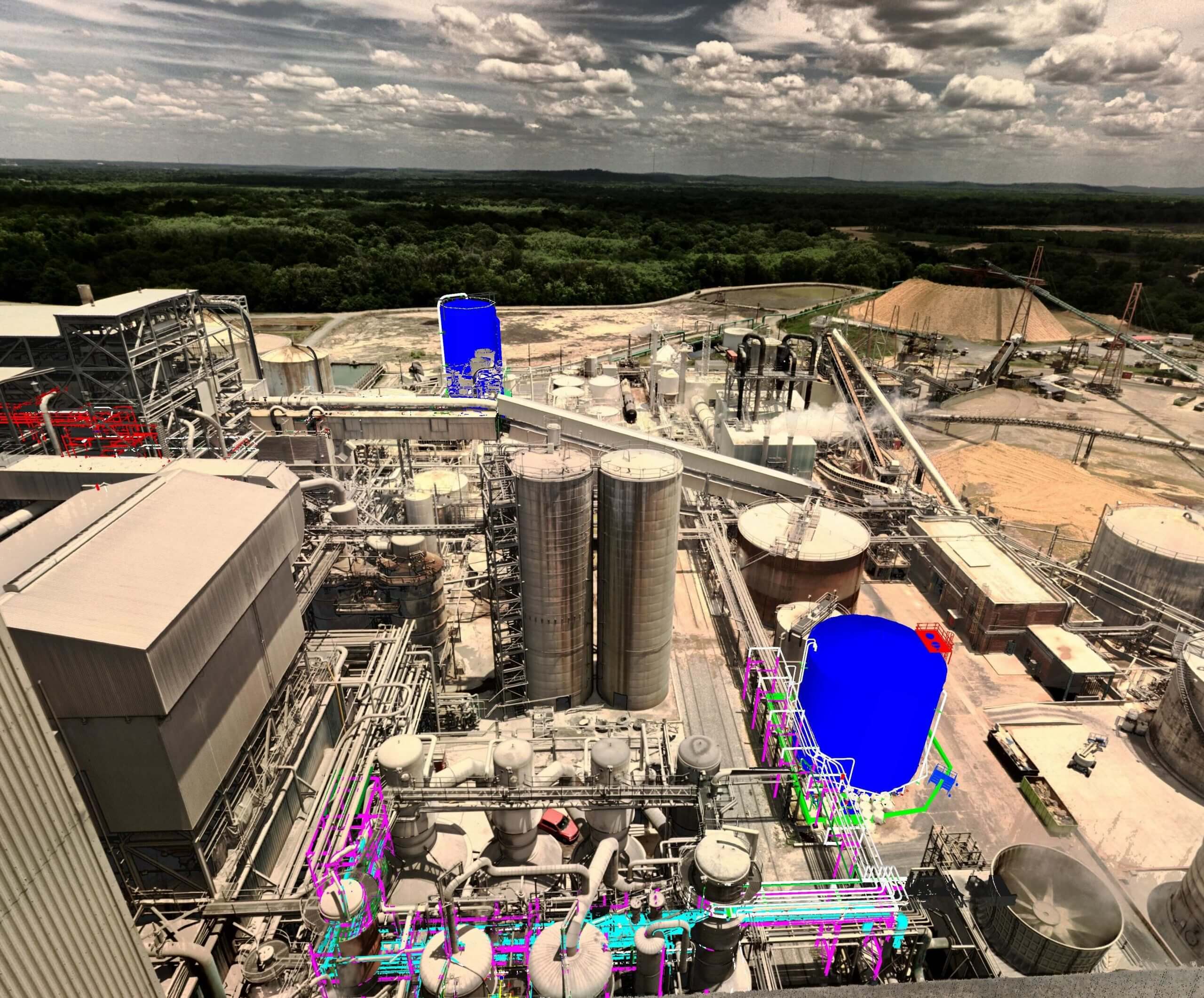 Aerial view of a paper facility featuring two large tanks, highlighted in blue, amidst complex industrial infrastructure. The landscape includes various pipes and structures under a cloudy sky.