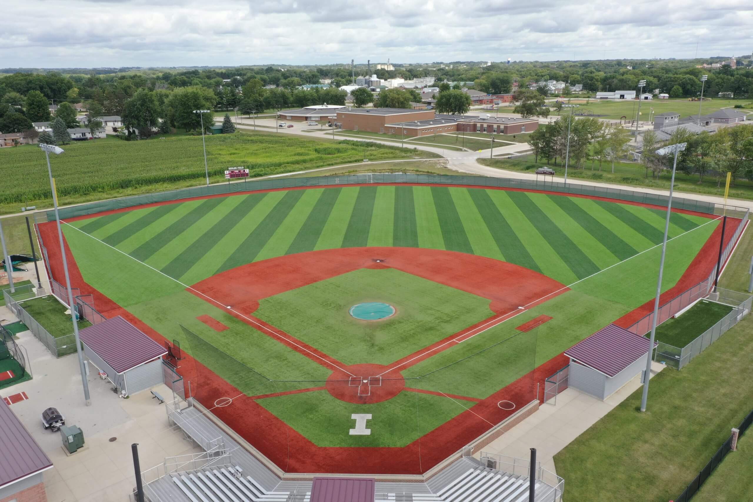 Aerial view of a newly improved baseball field with a turf infield and grass outfield, featuring clear striping patterns, surrounded by bleachers and adjacent facilities.