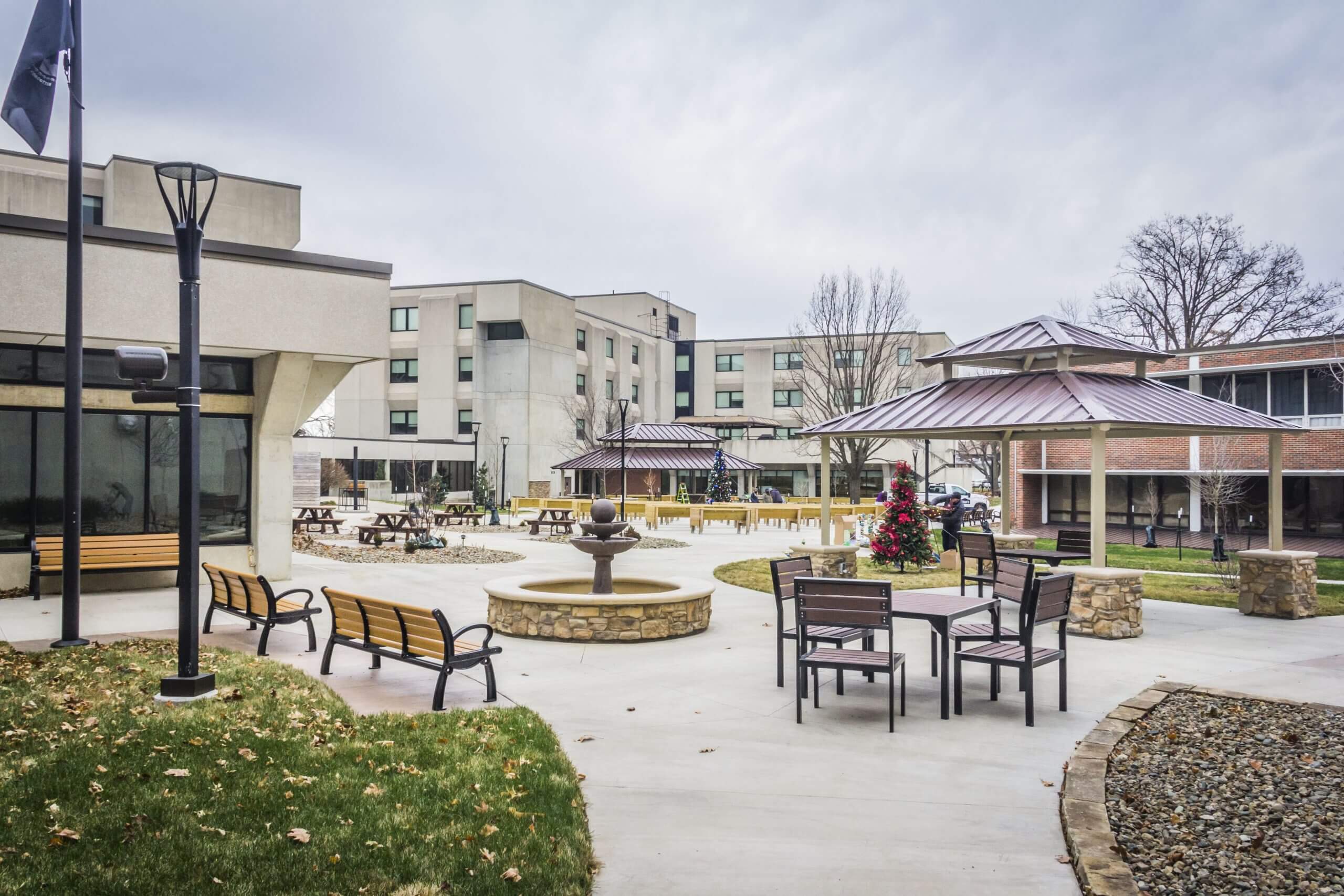 Courtyard at the Iowa Veterans Home featuring a central fountain, benches, tables, a gazebo, and surrounding buildings, with overcast skies.