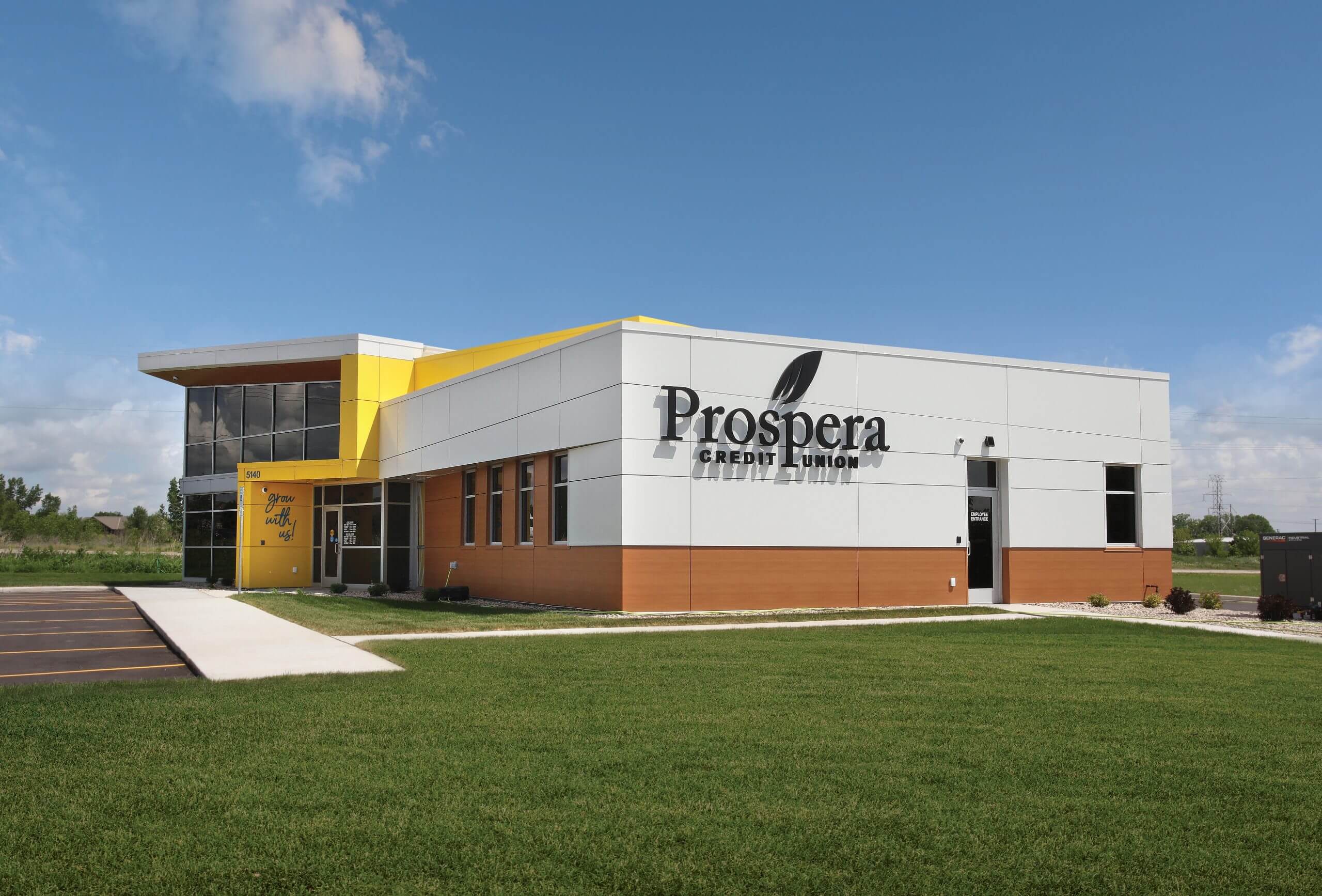 Prospera Credit Union building, a modern one-story structure with large glass windows, a yellow accent, and "Prospera Credit Union" text on the facade, set against a clear blue sky.