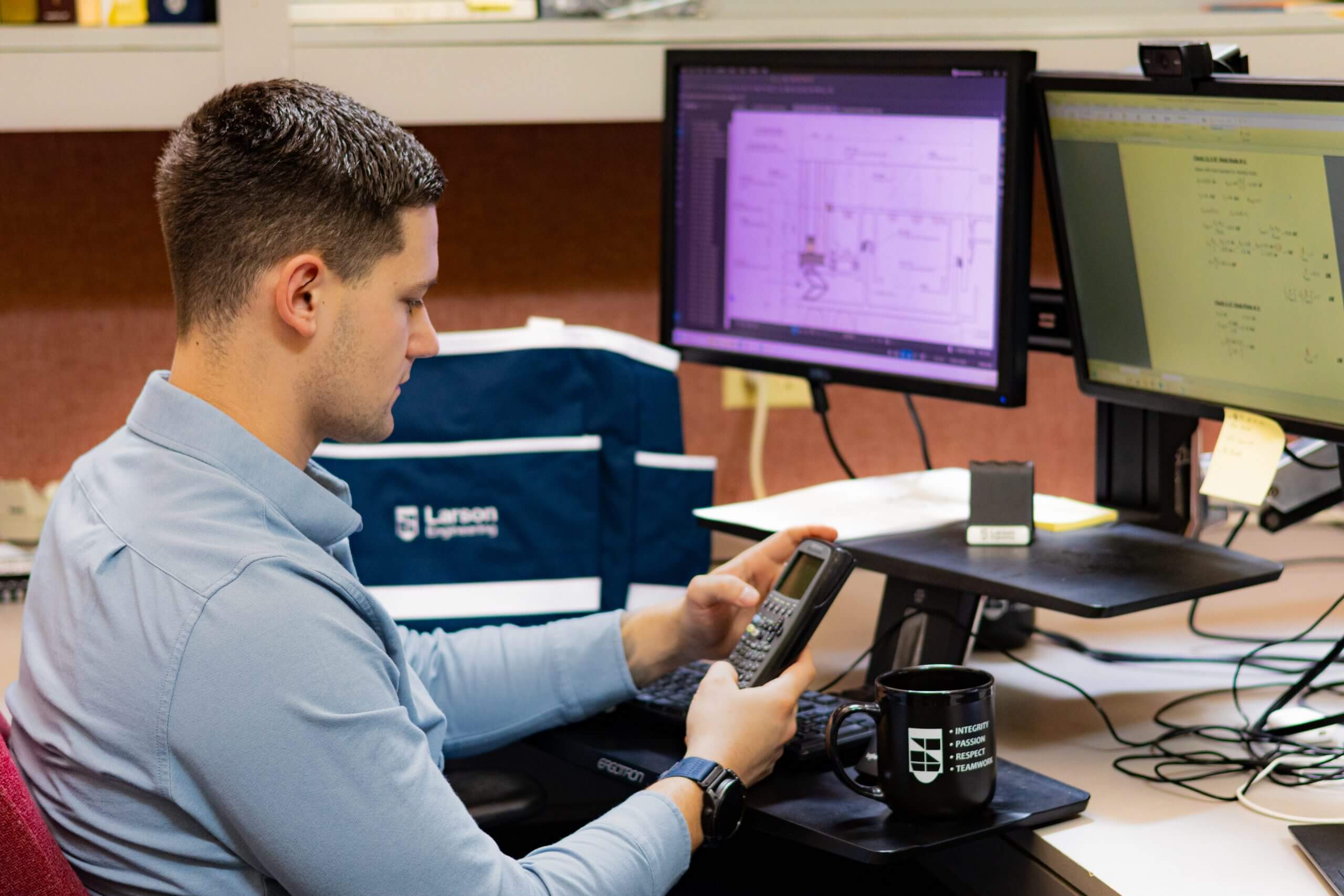 Engineer at a desk using a calculator, with two computer monitors displaying technical drawings and a Larson Engineering branded bag and mug visible.