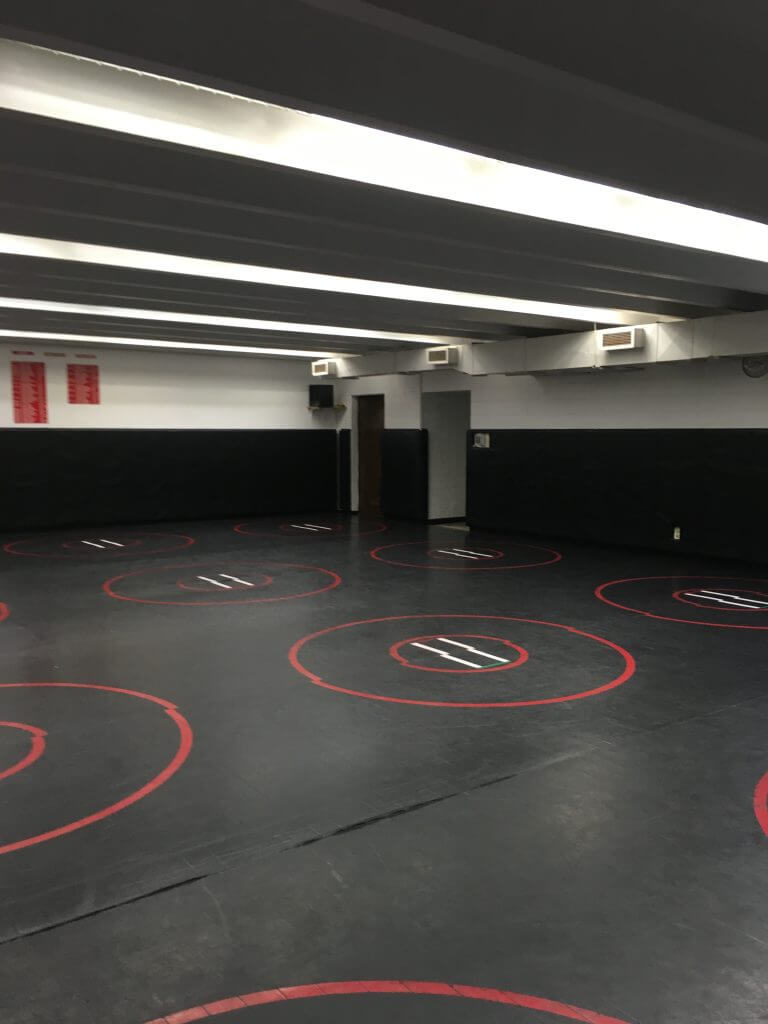 Wrestling room with black mats featuring red circles and lines, surrounded by padded walls and overhead lighting.