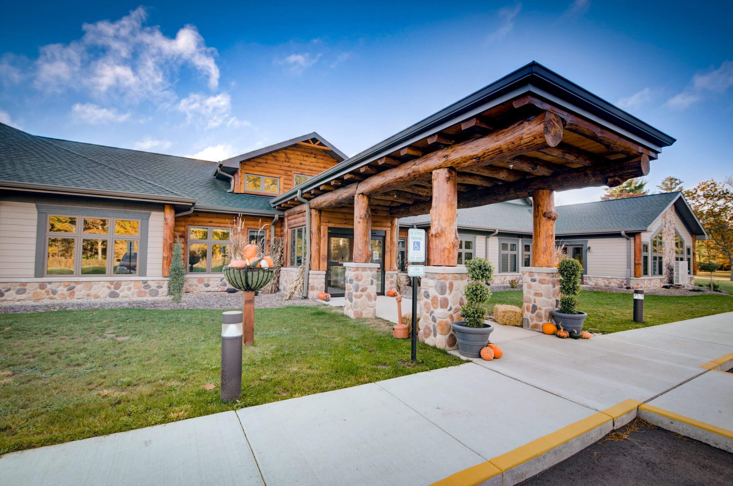 Riverwood Eagle’s Nest, a one-story wood-framed memory care and assisted living facility in Wisconsin Dells, WI, featuring a stone and wood exterior with a covered entrance adorned with potted plants and pumpkins.