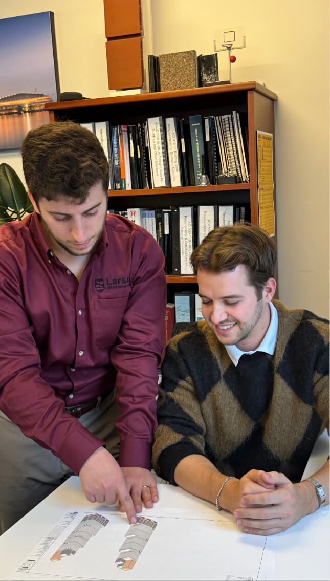 Two professionals at Larson Engineering reviewing architectural plans on a table, with one pointing at the drawings and the other observing, in an office setting with bookshelves in the background.